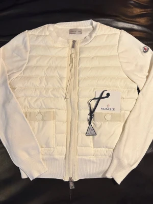 Moncler cardigan  - Snygg vit cardigan jacka från Moncler med quiltad framsida och stickade ärmar. Jackan har rund hals, två fickor fram och dragkedja hela vägen. Perfekt för dig som vill ha en stilren look med exklusiv känsla.