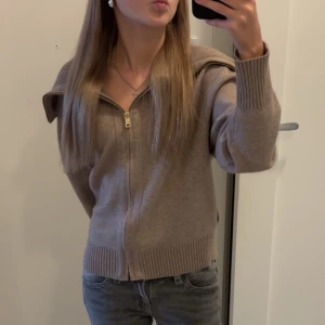 Beige stickad kofta med dragkedja - Säljer en beige stickad kofta med dragkedja framtill och breda ribbade muddar vid ärmslut och nederkant. Koftan har långärmad design och en snygg krage som ger en chill och trendig look. Perfekt att slänga på sig när det är lite kyligt ute.