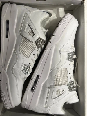 Nike Air Jordan 4 Retro "Pure Money - Nike Air Jordan 4 sneakers i helvit med snygga silverdetaljer vid snörningen. Skorna har meshpaneler på sidorna, klassisk snörning och en platt sula med Air-enhet. Materialet är skinn och mesh, och de har en rund tå. Kommer med originalkartong.