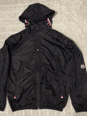 Svart vindjacka från Moncler med huva - Svart vindjacka från Moncler med huva och dragkedja framtill. Jackan har Moncler-logga på ärmen och snygga rödvita detaljer vid huvan och fickorna. Materialet är lätt och glansigt, perfekt för blåsiga dagar. Jackan har elastiska muddar och en clean, sportig look. Pris kan diskuteras 