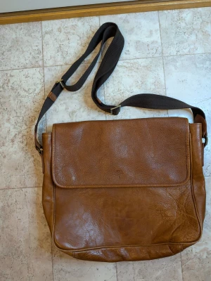 Brun axelväska från Mulberry i skinn - Säljer en snygg brun axelväska från Mulberry i äkta skinn. Väskan har ett klassiskt, stilrent utseende med justerbar axelrem i svart textil och praktiskt lock. Insidan har Mulberrys ikoniska trädmönster och en dragkedja med logga. Perfekt för dig som gillar tidlös design. Inte äkta.