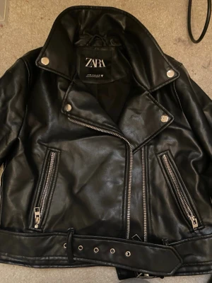 Svart skinnjacka från Zara - Säljer en svart skinnjacka från Zara med coola silvriga dragkedjor, bälte i midjan och klassisk bikerstil. Jackan har stora slag, långa ärmar och flera fickor med dragkedja. Perfekt för dig som vill ha en edgy look.