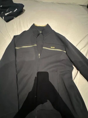 Svart ziptröja från BOSS XXL - Svart långärmad ziptröja från BOSS i storlek XXL. Tröjan har gul logga på bröstet och gul dekorativ linje över bröstet. Hög krage och dragkedja framtill. Snygg detalj med BOSS-band på insidan av kragen. Perfekt för en stilren och sportig look.