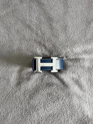 Hermès bälte  - Säljer ett stilrent Hermès skärp med klassiskt H-spänne i silverfärg. Skärpet är vändbart med ena sidan i mörkblått och andra i svart, tillverkat i äkta skinn med vit kontrastsöm. Perfekt accessoar för att lyfta din outfit.