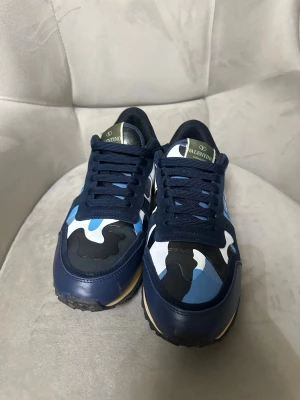Valentino Rockrunner sneakers blå camo - Säljer ett par Valentino Rockrunner sneakers med blått, svart och vitt camouflage-mönster. Skorna har snörning, rund tå och platt sula. Materialmix med mocka, skinn och textil samt detaljer med nitar på hälen. Snygg kontrast med beige mellansula och svart yttersula.