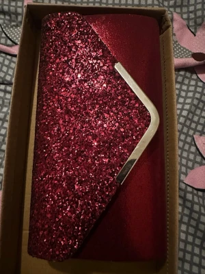 Röd glittrig kuvertväska - Snygg röd kuvertväska med glittrig framsida och metallkant. Väskan har en elegant och stilren design, perfekt för att lyfta din outfit. Materialet är syntet med massor av glitter som verkligen fångar ljuset.