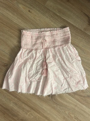 Ljusrosa kortkjol från Gina Tricot - Supersöt ljusrosa kortkjol från Gina Tricot med smockad midja och knytband med tofsar. Kjolen har ett diskret mönster och är gjord i mjuk bomull. Perfekt för dig som gillar en feminin och chill stil.