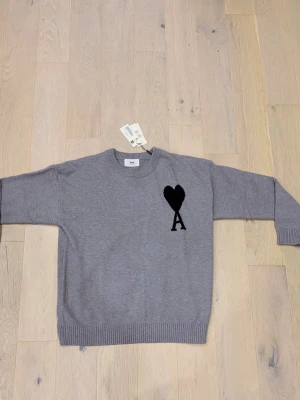 Grå stickad tröja från Ami Paris - Snygg grå stickad tröja från Ami Paris med svart hjärta och bokstaven A broderad på bröstet. Tröjan har rund hals, ribbade muddar och lång ärm. Perfekt för dig som gillar stilrena detaljer och vill ha något som sticker ut lite.