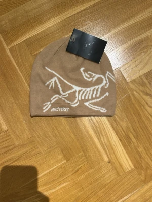 Beige mössa från Arc'teryx - Snygg beige mössa från Arc'teryx med vit dinosaurie-tryck på framsidan och logga. Mössan är mjuk och skön, perfekt för kalla dagar. Enkel design med stretchig passform som sitter bra på huvudet.