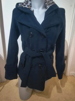 Mörkblå skepparkavaj med leopardfoder - Snygg mörkblå skepparkavaj med dubbelknäppning och bälte i midjan. Insidan av kragen och huvan har leopardmönstrat foder som ger en cool kontrast. Jackan har långa ärmar och stora knappar framtill. Perfekt för dig som gillar klassisk stil med en twist. Storlek 40