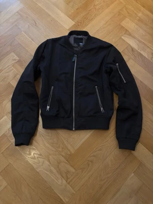 Svart bomberjacka stl 160 - Snygg svart bomberjacka med klassisk ribbad krage och muddar. Jackan har silverfärgade dragkedjor på fickorna och ärmen, samt en rak passform som ger en cool look. Perfekt för dig som gillar streetstyle och vill ha en enkel men stilren jacka.