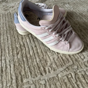 Adidas Campus rosa mocka sneakers - Säljer ett par Adidas Campus sneakers i ljusrosa mocka med vita ränder på sidorna och vit sula. Skorna har snörning och en rund tå, samt en gråblå detalj vid hälen. Perfekta för dig som gillar klassisk streetstil med en soft färg.