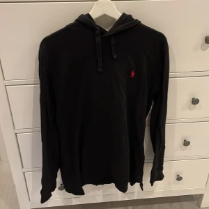 Svart hoodie från Polo Ralph Lauren - Svart hoodie från Polo Ralph Lauren med klassisk huva och snörning. Tröjan har lång ärm och en liten röd broderad logga på bröstet. Perfekt för en chill och stilren look.