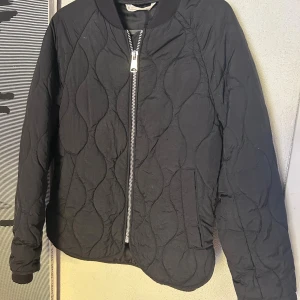 Svart quiltad bomberjacka H&M - Quiltad svart bomberjacka från H&M med snyggt mönster och silverfärgad dragkedja framtill. Jackan har ribbad krage och muddar, samt två fickor på sidorna. Perfekt för dig som gillar stilren och enkel design.