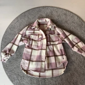 Rutig kappa i rosa och beige - Säljer en mysig kappa från med rutigt mönster i ljusrosa, beige och vitt. Kappan har två stora bröstfickor med lock, klassisk krage och knappar framtill. Perfekt för lager-på-lager och chill stil.