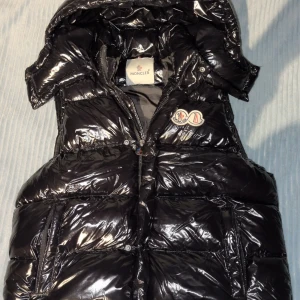 Hittar it bild på Burberry västen  - Snygg svart pufferjacka från Moncler med glansig finish och huva. Jackan har dragkedja framtill, två sidofickor med dragkedja och klassiska Moncler-loggor på bröstet. Perfekt för kalla dagar när du vill hålla stilen.