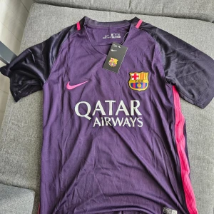 FC Barcelona Lamine Yamal tröja Nike - Säljer en officiell FC Barcelona matchtröja från Nike med Lamine Yamal och nummer 10 på ryggen. Tröjan är lila med svarta ärmar, rosa detaljer och klubbmärke på bröstet. Tillverkad i lätt och ventilerande polyester med Dri-FIT-teknologi.