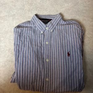 Randig blå skjorta från Polo Ralph Lauren - Snygg blå skjorta från Polo Ralph Lauren med smal passform och klassisk krage. Skjortan har vita och röda vertikala ränder, knappar framtill och det ikoniska broderade Polo-logot på bröstet. Perfekt för dig som gillar stilrena detaljer och tidlös design.