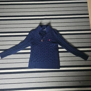 Marinblå stickad tröja Polo Ralph Lauren - Säljer en marinblå kabelstickad tröja från Polo Ralph Lauren med halv dragkedja och klassisk krage. Tröjan har lång ärm, ribbade muddar och en liten röd broderad logga på bröstet. Perfekt för lager-på-lager och snygg till jeans.