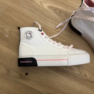 Hello Kitty vita high top sneakers - Snygga vita high top sneakers med rosa och svarta detaljer från Hello Kitty. Skorna har rosa/vita snören, svart hälparti med Hello Kitty-tryck och en söt Hello Kitty-logga på sidan. Insidan är rosa och materialet är syntet. Perfekt för dig som vill sticka ut med en gullig och cool stil.