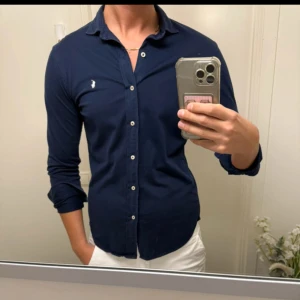 Mörkblå skjorta från Polo Ralph Lauren - Snygg mörkblå skjorta från Polo Ralph Lauren med klassisk krage och vita knappar. Skjortan har lång ärm och ikonisk broderad logga på bröstet. Tillverkad i mjuk bomull som känns skön mot huden. Perfekt för dig som gillar stilrena och tidlösa plagg.