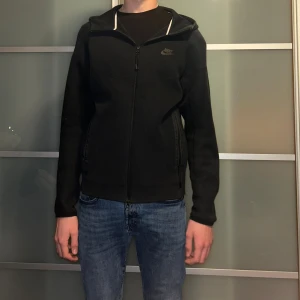 Svart Nike tech fleece - Svart hoodiejacka från Nike med dragkedja framtill och två sidofickor. Jackan har en stilren design med Nike-logga på bröstet och huva med vit kant. Perfekt för chill dagar och streetwear-stil.