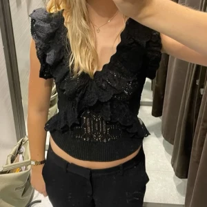 Svart spetsig bolerotopp med volanger - Säljer en svart bolerotopp från H&M med spetsdetaljer och volanger längs halsringningen och axlarna. Toppen har en omlottkänsla och är tillverkad i ett mjukt, stickat material med dekorativ spets. Perfekt för dig som gillar romantiska vibes och snygga detaljer.