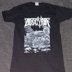 Svart Obscura band t-shirt - Säljer en svart t-shirt med tryck från metalbandet Obscura. Framsidan har ett stort vitt bandlogo-tryck och detaljerad grafik med dödskallar och landskap. Baksidan har ett tryck med en korp på ett kors och datumet 1.11.2010. Perfekt för dig som gillar metal och unika bandtröjor.