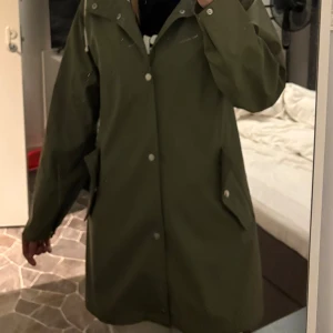 Regnjacka - Säljer en olivgrön regnjacka med huva och tryckknappar framtill.