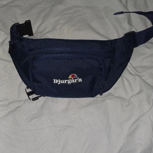 Blå Djurgår'n midjeväska - Snygg marinblå midjeväska från Djurgår'n med broderad logga framtill. Väskan har flera fack med dragkedja och justerbart band med spänne. Perfekt för att bära med sig det viktigaste på stan eller till matchen.