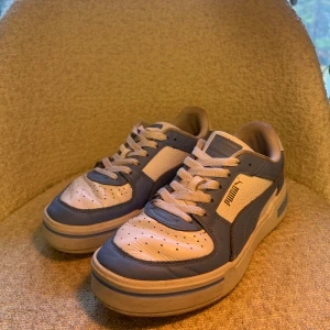 Puma sneakers i blått och vitt, strl 39 - Snygga Puma sneakers i ljusblått och vitt med klassisk logga på sidan och hälen. Skorna har snörning, perforerad tå och platt sula. Tillverkade i skinn och syntet med rund tå. Perfekt för dig som gillar sportig stil och vill ha något som sticker ut.