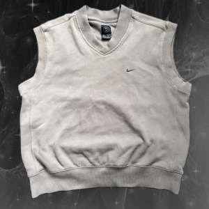 Beige väst från Nike med v-ringning - Snygg beige väst från Nike med v-ringad hals och diskret broderad logga på bröstet. Västen är i mjuk bomull och har ribbade kanter vid hals och midja. Perfekt att slänga över en t-shirt för en chill look.