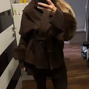 Brun kappa med pälsdetaljer och bälte - Säljer en brun kappa med bred krage och pälsdetaljer vid ärmsluten. Jackan har ett bälte i midjan för snygg passform och är perfekt för kyliga dagar. Stilen är loose och den har långa ärmar med stickade muddar.