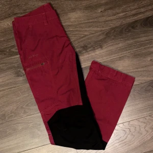 Lundhags röda friluftsbyxor AVHU - Snygga vinröda friluftsbyxor från Lundhags, modell AVHU. Byxorna har svarta förstärkningar på knän och bak, praktiska fickor med dragkedja och knappdetaljer vid benslut. Perfekta för äventyr och outdoor, designade i slitstarkt material. Använda fåtal gånger så i fint skick! (Öppen för prisförslag)