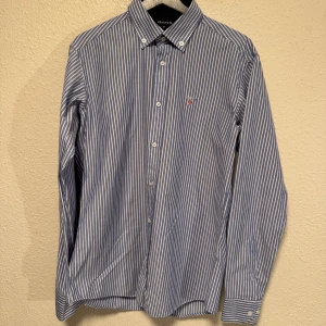 Blåvit randig skjorta från Gant M - Klassisk blå och vit randig skjorta från Gant med button down-krage och broderad logga på bröstet. Skjortan har långa ärmar, knappar framtill och är tillverkad i mjuk bomull. Perfekt för en clean och stilren look.