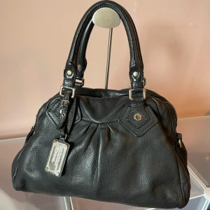 Svart handväska från Marc by Marc Jacobs - Snygg svart handväska från Marc by Marc Jacobs i äkta skinn. Väskan har två rejäla handtag, silverfärgade metalldetaljer och en cool logotypbricka framtill. Insidan är fodrad med mönstrat tyg och har dragkedja upptill. Perfekt för dig som gillar klassisk stil med edge.