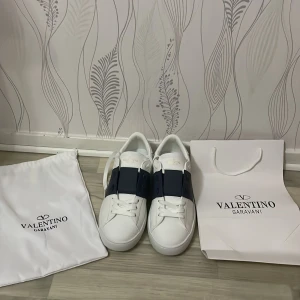 Valentino Garavani Open sneakers vit/blå - Säljer ett par lyxiga Valentino Garavani Open sneakers i vit kalvskinn med bred mörkblå rem över ovansidan. Snygg och stilren design med vita snören och subtila nitar på hälen. Kommer med originalpåse och låda. Perfekt för dig som gillar exklusiva sneakers. Priset går att diskutera 