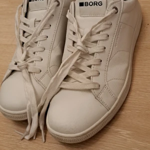 Björn Borg vita sneakers med svart detalj - Fräscha vita sneakers från Björn Borg med svart hälparti och klassisk snörning. Skorna har rund tå, platt sula och är tillverkade i skinn med diskreta sömmar och logotyp på sidan och bak. Perfekt för dig som gillar stilrena och cleana sneakers.