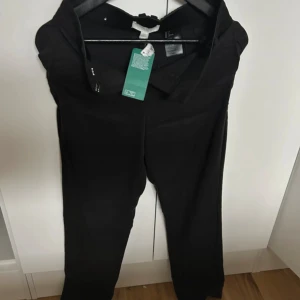 Svarta kostymbyxor från H&M - Stilrena svarta kostymbyxor från H&M med klassisk rak passform. Byxorna har hällor för bälte och stängs med dragkedja och knapp. Perfekta för en clean och dressad look.