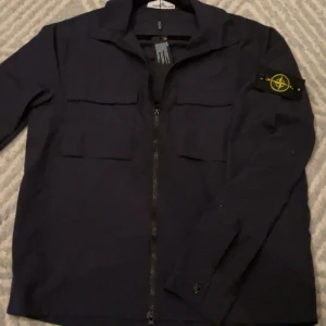 Mörkblå overshirt från Stone Island - Säljer en mörkblå overshirt från Stone Island med klassisk kompasspatch på ärmen. Jackan har två bröstfickor med lock, hel dragkedja framtill och en stilren krage. Perfekt för dig som gillar vill klä dig presentabelt nu till höst!!! 🫡🔥