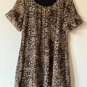 Leopardmönstrad kortklänning med volang - Snygg kortklänning med leopardmönster i beige och bruna toner. Klänningen har volangdetaljer på ärmar och nederkant, samt en rund halsringning. Materialet är lätt och transparent, perfekt för dig som vill sticka ut med ett djuriskt print.