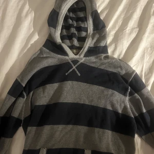 Randig hoodie från Dissident - Snygg hoodie från Dissident med breda mörkblå och grå ränder. Tröjan har huva med ribbad kant och klassisk känguruficka framtill. Perfekt för chill dagar och har en avslappnad passform. Materialet känns mjukt och bekvämt mot huden.