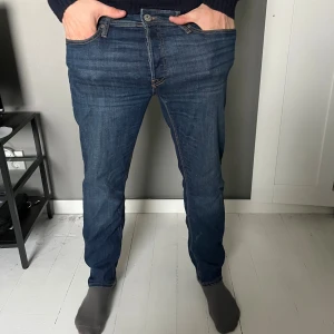 Jack&jones  mörkblå jeans - Snygga jacknjones jeans i mörkblå tvätt med klassiska fem fickor och bruna sömmar. Modellen är slim och har rak passform med normal midja. Jeansen är tillverkade i slitstarkt denim och har en läderpatch bak i midjan.