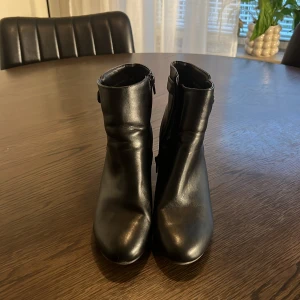 Svarta stövletter från Anna Field - Snygga svarta stövletter från Anna Field i skinnimitation med rund tå och blockklack. Skorna har dragkedja på sidan och en dekorativ rem med metallknapp runt ankeln. Perfekt till höstens och vinterns outfits.