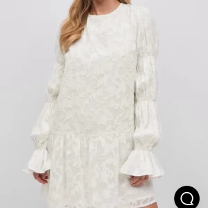 Studentklänning - Superfin vit kort klänning med subtilt blommönster och volangdetaljer på ärmarna. Klänningen har långärmade puffärmar med smock och en lös passform. Perfekt för dig som gillar romantisk och trendig stil.