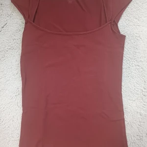 Vinröd topp från Brandy Melville - Vinröd enkel topp från Brandy Melville med rund halsringning och korta ärmar. Modellen är figurnära och tillverkad i mjuk bomull med lite stretch för en bekväm passform. Perfekt att styla med jeans eller kjol för en clean look.