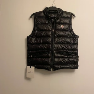 Svart dunväst från Moncler/Moncler gui väst  - Svart dunväst från Moncler med quiltad design och glansig finish. Västen har två sidofickor med dragkedja, dubbel dragkedja framtill och Moncler-logga på bröstet. Perfekt för lager-på-lager och snygg till streetwear. Skick 10/10 använd endast en gång 