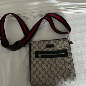  Axelväska med GG-mönster - Snygg axelväska med klassiskt GG-mönster i brunt på beige bakgrund. Väskan har svart läderdetalj runt dragkedjan framtill och en bred axelrem i marinblått och rött. Perfekt för dig som vill ha en stilren och lyxig accessoar.