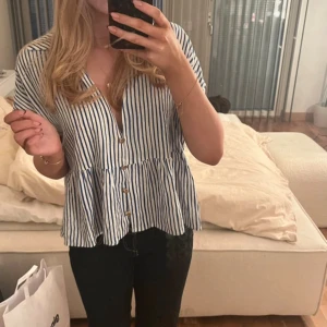 Randig blus från Zara - Säljer en fräsch blus från Zara i storlek S. Den är vit med blå ränder, har v-ringning, korta ärmar och knappar framtill. Blusen har en volang nedtill som ger en chill och flowy vibe. Materialet är mjuk bomull, perfekt för varma dagar.