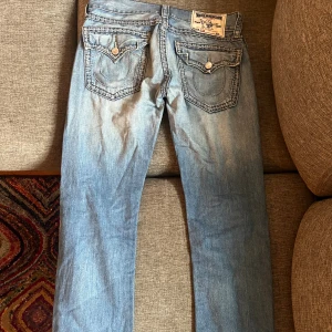 True Religion ljusblå jeans med detaljer - Säljer ett par ljusblå jeans från True Religion med snygga slitningar och kontrastsömmar. Modellen har klassiska fickor med lock och silverfärgade knappar, samt logopatch bak. Jeansen har rak passform och är tillverkade i jeansmaterial. Längd skulle säga 30 och midja ish 31.